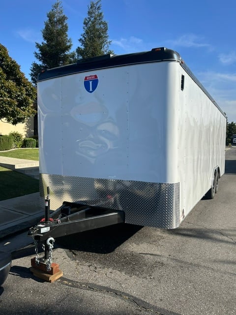 White enclosed cargo trailer with Interstate shield logo and hitch connection parked on asphalt with trees in background